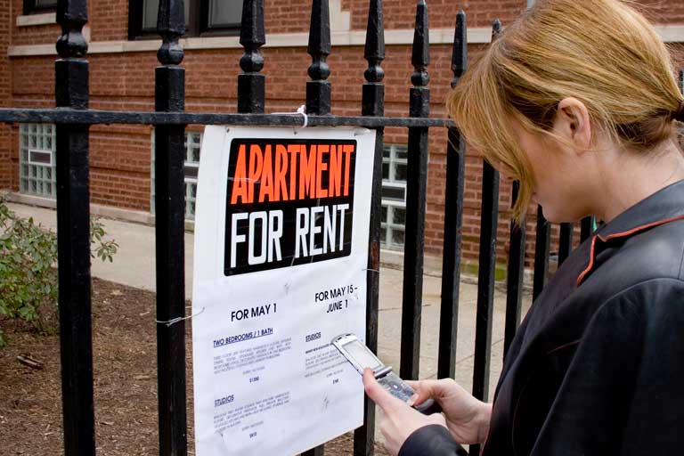 Woman standing in front of a gated brick building. Sign on the gate says "Apartment for rent.".