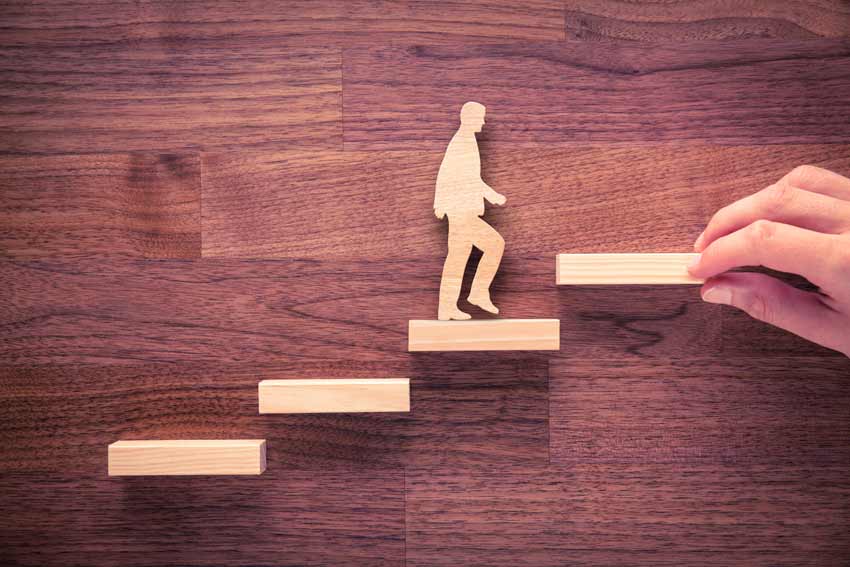 wooden figure of a man stepping up wood blocks; the final step is held by a human hand.