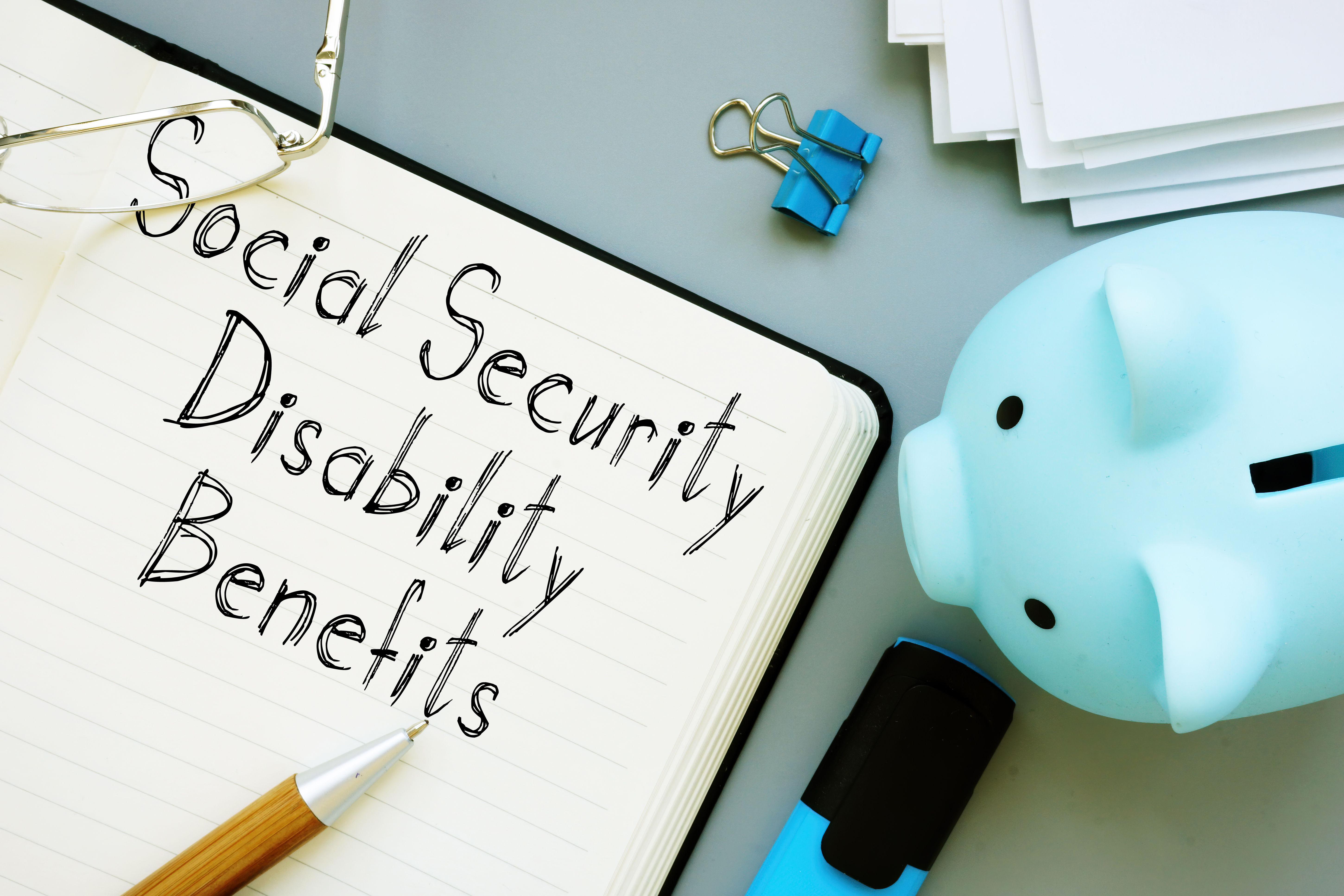 Notebook with the words Social Security Benefits Information. Next to the notebook is a small blue piggy bank.