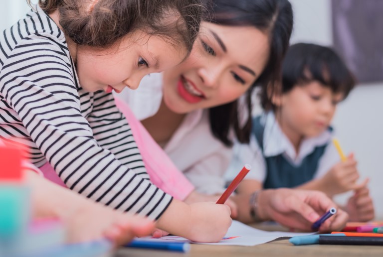 A preschool teacher talks with a young girl while she is drawing.